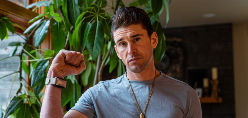 Ben Greenfield raises a fist toward the camera while looking directly at the viewer with a serious expression. He wears a gray t-shirt, a gold pendant necklace, and a smartwatch. A large green leafy plant and a warmly lit interior are visible in the background.