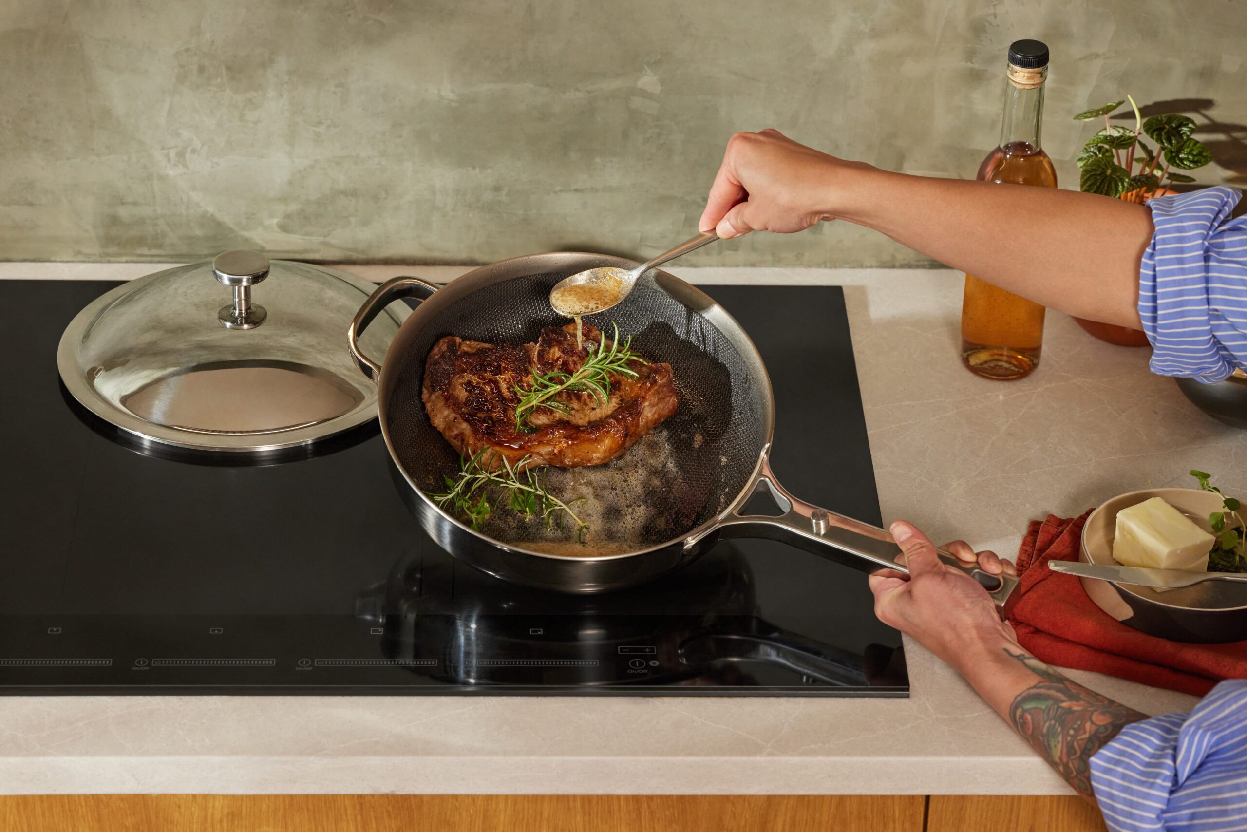A person basting a seared steak with a spoon over a stainless steel Our Place pan on a black induction cooktop. The steak is surrounded by fresh rosemary and thyme sprigs in bubbling butter. The pan's matching lid rests on the counter to the left. In the background, a bottle of amber liquid, a small green plant, and a small dish of butter with herbs are visible on a light stone countertop.