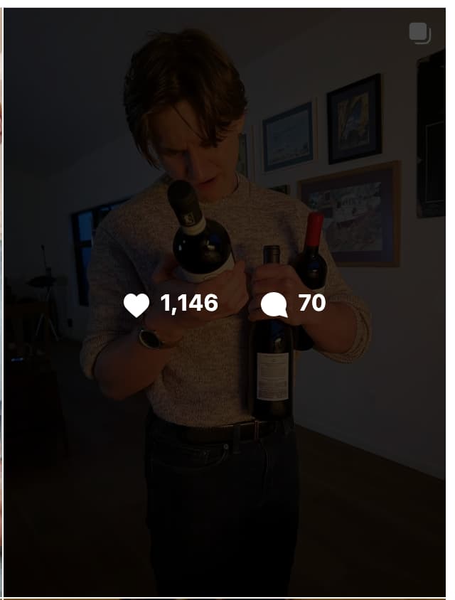 A young man with brown hair wearing a light gray long-sleeve sweater and dark pants stands indoors in a dimly lit room, holding three wine bottles cradled in both arms and looking down at them. He is wearing a dark watch on his left wrist. In the background, several framed pictures hang on a white wall, and a lamp or light stand is visible to the left. A social media engagement overlay shows 1,146 likes and 70 comments at the center of the image.