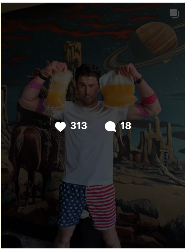 A man stands indoors holding two clear medical IV fluid bags above his head, one in each raised hand. He is wearing a white t-shirt and American flag-patterned board shorts featuring red, white, and blue stars and stripes. Pink medical bands are visible on both of his upper arms, consistent with IV therapy use. He has a serious, intense expression on his face and is wearing a dark watch on his left wrist. The background features a large, colorful painted mural depicting a surreal Western-meets-outer-space scene, with a ringed planet in the upper right, red rock desert formations, a night sky with stars, and a figure on horseback visible on the left side. A social media engagement overlay shows 313 likes and 18 comments.