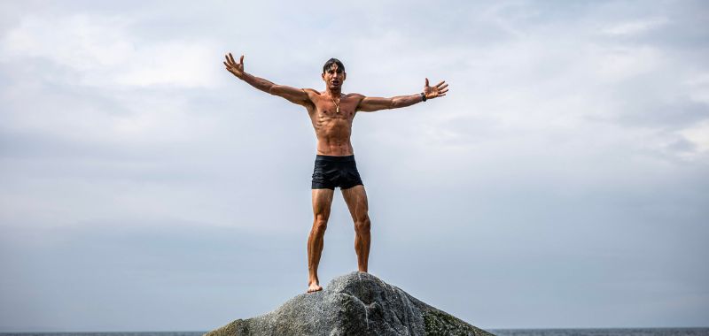 Ben Greenfield a lean, muscular shirtless man wearing black swim shorts stands atop a large rocky outcropping with his arms spread wide open in a triumphant pose, facing the camera. He is barefoot and wearing a watch and a necklace. The background features a dramatic overcast sky with thick gray clouds and the ocean horizon visible behind him. The wide-angle shot is taken from below, emphasizing his silhouette against the sky.