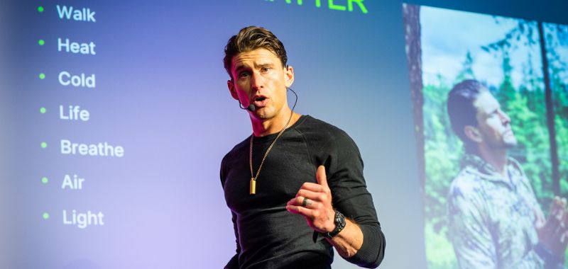 Ben Greenfield speaking on stage with a headset microphone and gold pendant necklace, gesturing while presenting in front of a projected slide listing wellness practices including Walk, Heat, Cold, Life, Breathe, Air, and Light.