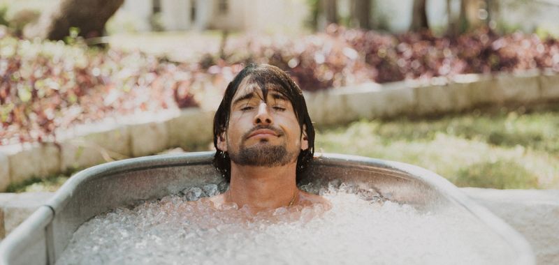 A man with long dark hair and a beard submerged in an outdoor ice bath, eyes closed and expression calm, with greenery and a garden wall visible in the background.