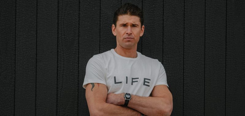 Ben Greenfield standing in front of dark background wearing a white shirt that says "LIFE"