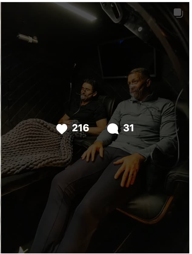 Video thumbnail of two men reclining in leather chairs inside a hyperbaric oxygen chamber, both wearing oxygen masks, with 216 likes and 31 comments.