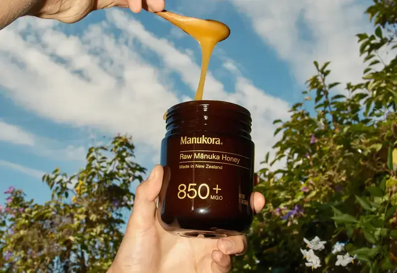 Hand holding a jar of Manukora Raw Manuka Honey MGO 850+ from New Zealand with honey dripping from a spoon against a blue sky and greenery background
