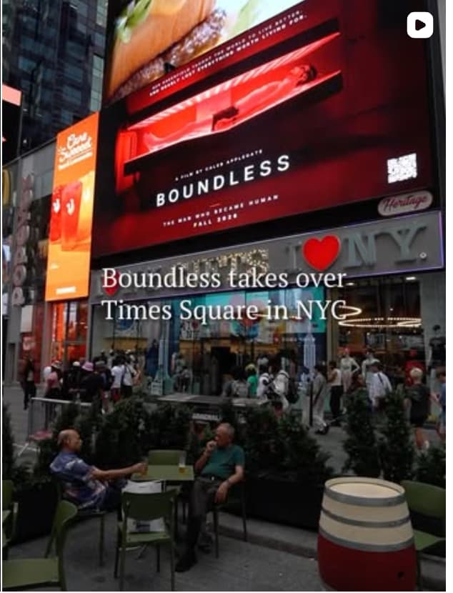 A large digital billboard in Times Square displays a poster for the film "Boundless: The Man Who Became Human," showing a person lying inside a red-lit therapy bed. Text overlaid on the photo reads "Boundless takes over Times Square in NYC." Crowds walk past an I Love NY gift shop below, and two men sit at an outdoor table in the foreground.