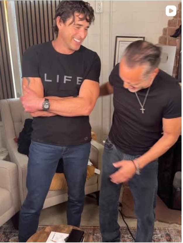 Two men in an indoor setting. The man on the left, wearing a black "LIFE" t-shirt with arms crossed, smiles at the camera. The man on the right in a black shirt stands beside him in a casual pose.