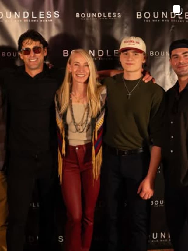 Four people pose together in front of a 'Boundless' branded backdrop. From left to right: a man wearing black sunglasses and dark clothing, a woman with blonde hair in a colorful patterned cardigan and red pants, a younger person in a dark shirt and cream-colored baseball cap with a cross necklace, and a man in dark clothing on the right.
