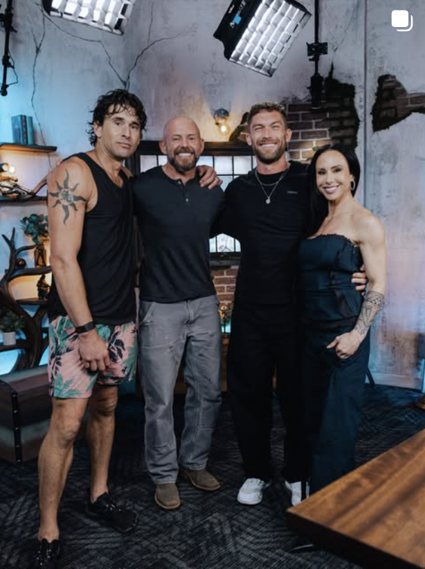 Group photo of four people posing arm-in-arm on a dimly lit studio set with exposed brick walls, string lights, and softbox lighting overhead. From left to right: a man with curly dark hair and a sun tattoo on his shoulder wearing a black tank top and tropical-print shorts; a bald man with a beard in a black button-down shirt and gray pants; a tall man with short hair in an all-black outfit; and a woman with long dark hair wearing a black off-shoulder jumpsuit.