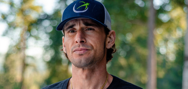 Portrait of a man with wavy shoulder-length brown hair, wearing a navy and white trucker hat with a green swoosh logo and a gold chain necklace over a dark shirt, standing outdoors against a blurred green forest background.