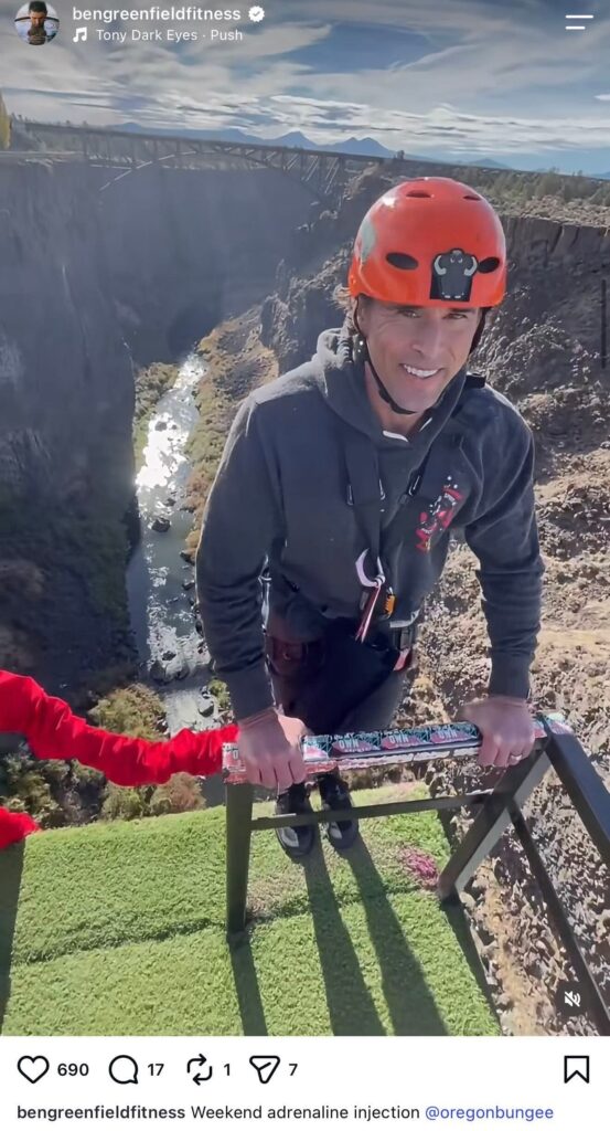 Instagram post from bengreenfieldfitness captioned ‘Weekend adrenaline injection’ and tagged @bungeebungee. Ben Greenfield, a man with short hair, wearing an orange climbing helmet and a gray jacket with a climbing/bungee harness, stands at a railing overlooking a deep canyon with a river and waterfalls below, with another person partially visible at the edge of the frame