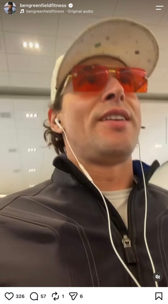 Instagram Reel from bengreenfieldfitness using original audio. Close-up selfie-style video still of Ben Greenfield, a man with wavy dark hair, wearing a dirty tan baseball cap, orange-tinted sunglasses, and a dark zip-up jacket with a white earbud cord visible, talking while looking upward in what appears to be an airport terminal.