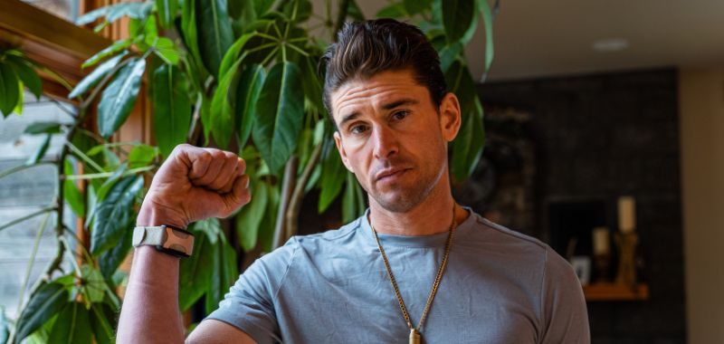 Ben Greenfield raises a fist toward the camera while looking directly at the viewer with a serious expression. He wears a gray t-shirt, a gold pendant necklace, and a smartwatch. A large green leafy plant and a warmly lit interior are visible in the background.