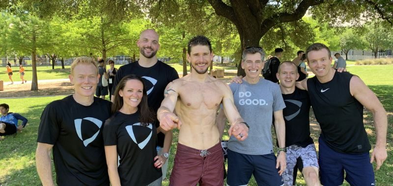 A group of seven people posing together outdoors in a sunny park with lush green trees in the background. From left to right: a lean man in a black t-shirt with a geometric logo, a smiling woman in a matching black logo t-shirt, a tall bald man in a black logo t-shirt standing behind the group, a shirtless man with a beard and a visible arm tattoo wearing dark red shorts standing in the center with arms outstretched, a man in a gray "Do Good." t-shirt, a shorter man in athletic wear, and a tall man in a black Nike sleeveless shirt. Most are smiling and appear to be in a post-workout or fitness event setting. Other people and park surroundings are visible in the background.