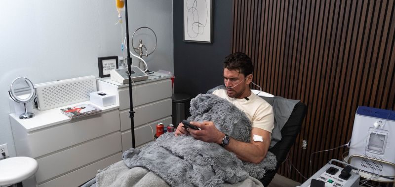 A man with dark hair and light stubble sits reclined in a clinical treatment chair in a modern, minimalist wellness clinic room. He is wearing a white t-shirt, a watch on his left wrist, and has a nasal cannula oxygen tube under his nose. He is looking down at a handheld device or phone held in both hands, with a large fluffy gray blanket draped over his lap and legs. An IV drip stand with a yellow-tinted fluid bag is visible to his upper left, with tubing connected to his arm. A medical monitoring device is positioned to his right.