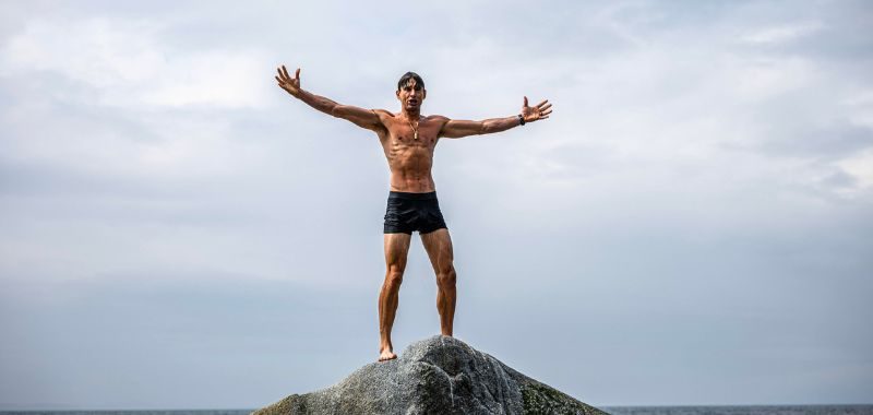 Ben Greenfield a lean, muscular shirtless man wearing black swim shorts stands atop a large rocky outcropping with his arms spread wide open in a triumphant pose, facing the camera. He is barefoot and wearing a watch and a necklace. The background features a dramatic overcast sky with thick gray clouds and the ocean horizon visible behind him. The wide-angle shot is taken from below, emphasizing his silhouette against the sky.