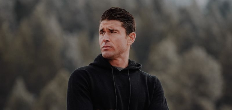 Ben Greenfield wearing a black hoodie with arms crossed, looking off to the side against a blurred outdoor background of trees and overcast sky.