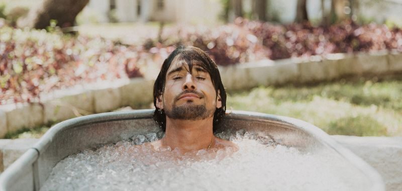 A man with long dark hair and a beard submerged in an outdoor ice bath, eyes closed and expression calm, with greenery and a garden wall visible in the background.