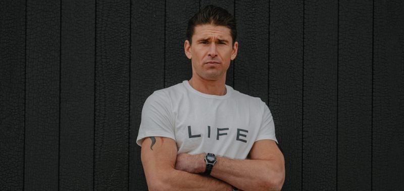 Ben Greenfield standing in front of dark background wearing a white shirt that says "LIFE"