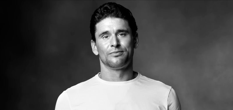 Black-and-white portrait of Ben Greenfield wearing a plain white t-shirt, looking directly at the camera with a neutral expression against a dark gray background.