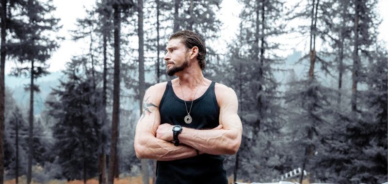 A man in a black sleeveless tank top stands outdoors with arms crossed, smiling at the camera. He's positioned in front of a misty forest with tall trees in the background, creating a moody, atmospheric setting.