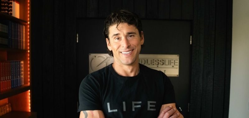 A man wearing a black "LIFE" t-shirt smiles at the camera in an indoor setting. Behind him is a doorway and warm orange-tinted lighting visible on the left side of the frame.