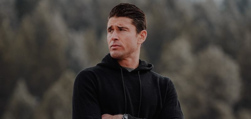 Portrait of a Ben Greenfield with short dark hair wearing a black hoodie, arms crossed, standing outdoors with a blurred backdrop of evergreen trees. He looks off to the side with a serious expression.